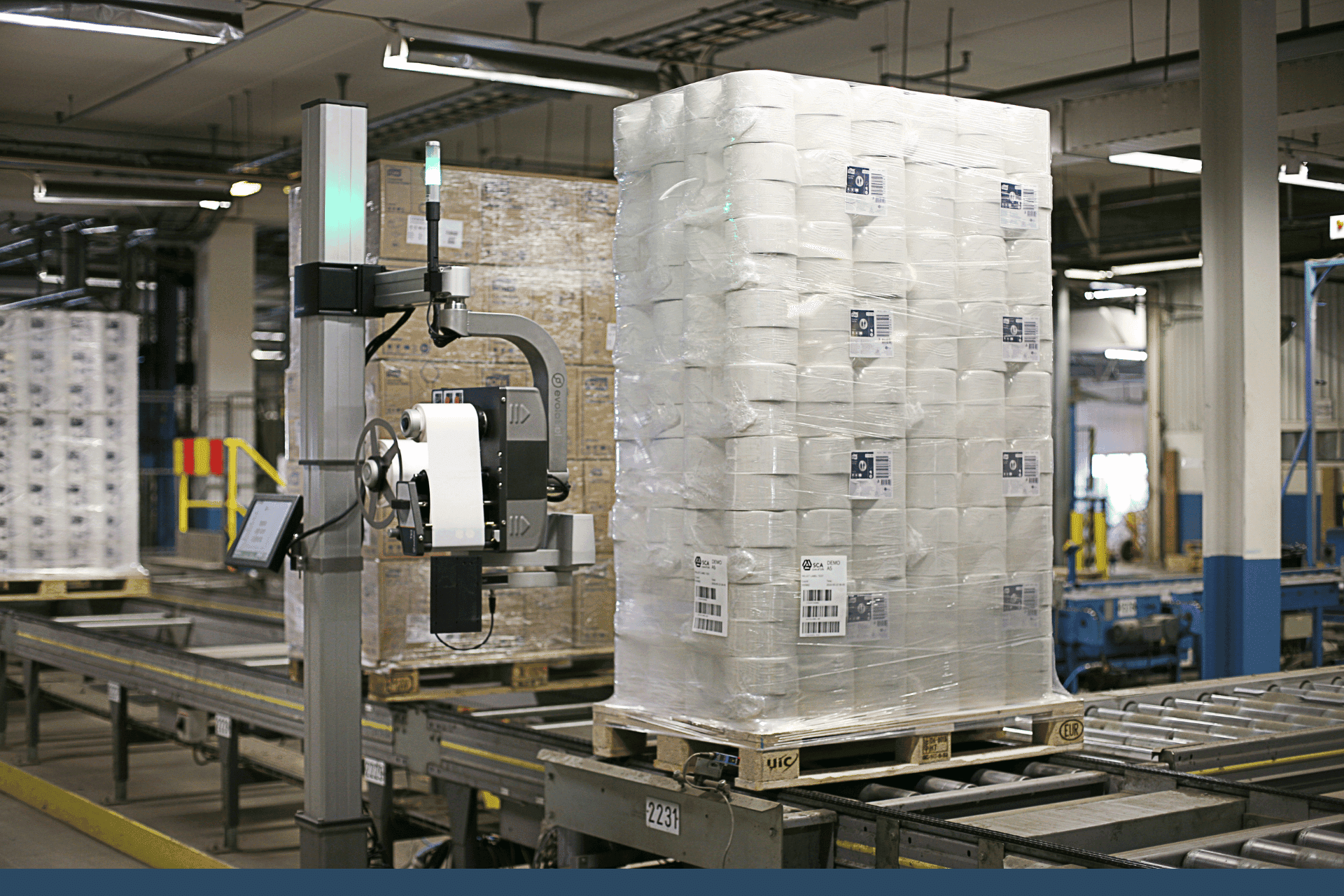 Paper rolls stacked on a pallet, on a production line with a labelling machine applying labels.